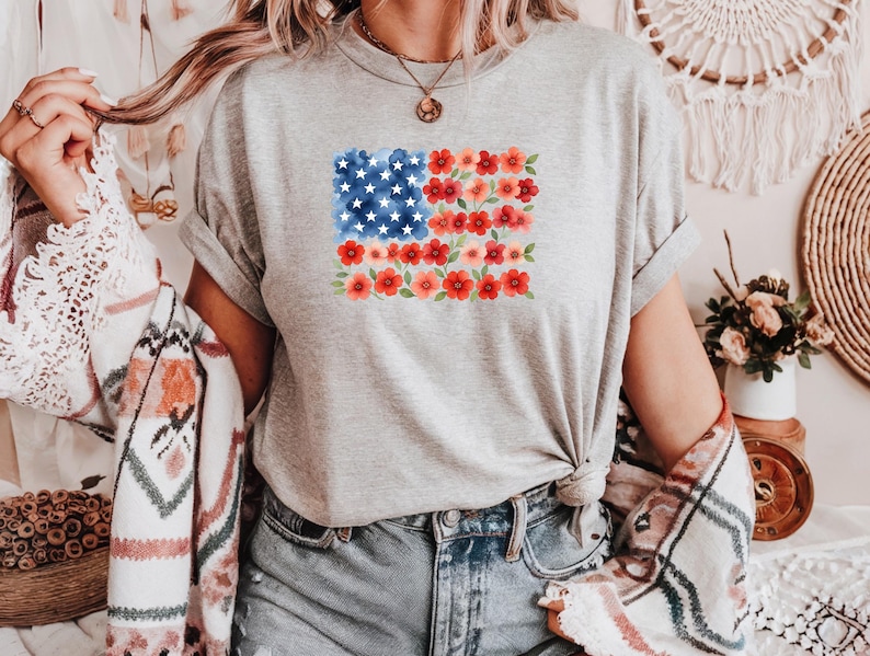 May include: A gray t-shirt featuring a watercolor design of the American flag. The flag design incorporates blue stars and red flowers. The shirt is paired with blue jeans and a patterned shawl.