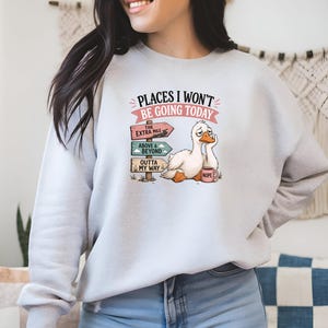May include: A light gray crewneck sweatshirt featuring a graphic of a tired white duck sitting next to a wooden signpost with three directional signs that read The Extra Mile, Above & Beyond, and Outta My Way, with the text Places I Won't Be Going Today.