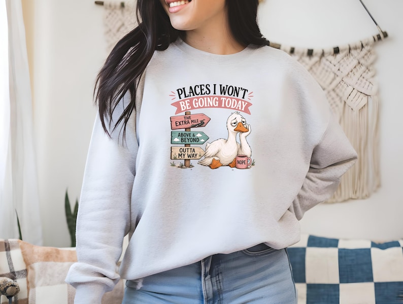 May include: A light gray crewneck sweatshirt featuring a graphic of a tired white duck sitting next to a wooden signpost with three directional signs that read The Extra Mile, Above & Beyond, and Outta My Way, with the text Places I Won't Be Going Today.
