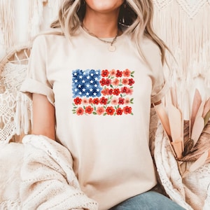 May include: A beige t-shirt featuring a floral American flag design. The flag is composed of blue watercolor stars, red flowers, and red and pink floral stripes. The shirt has rolled-up sleeves.