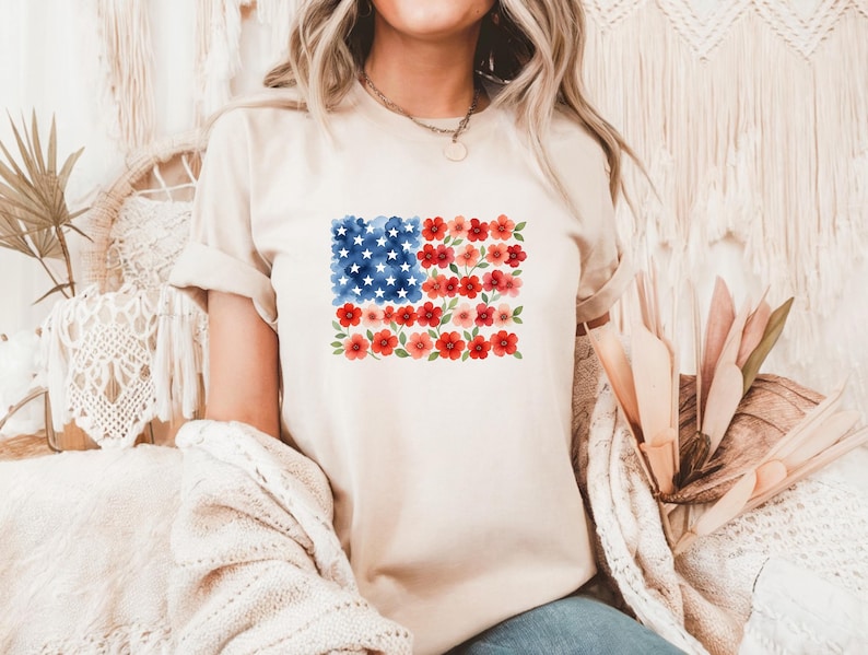 May include: A beige t-shirt featuring a floral American flag design. The flag is composed of blue watercolor stars, red flowers, and red and pink floral stripes. The shirt has rolled-up sleeves.
