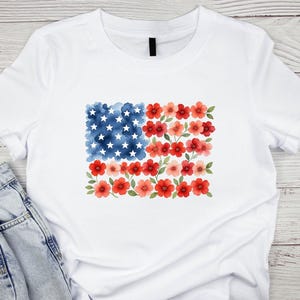 May include: White t-shirt featuring a watercolor design of the American flag. The flag design replaces the stripes with red flowers and the stars with white stars on a blue watercolor background. The shirt is made of a soft, comfortable material.