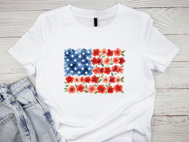 May include: White t-shirt featuring a watercolor design of the American flag. The flag design replaces the stripes with red flowers and the stars with white stars on a blue watercolor background. The shirt is made of a soft, comfortable material.