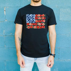 May include: Black t-shirt featuring a floral American flag design. The flag's stars are represented by white flowers on a blue background, and the stripes are red flowers. The shirt is a classic crew neck style.