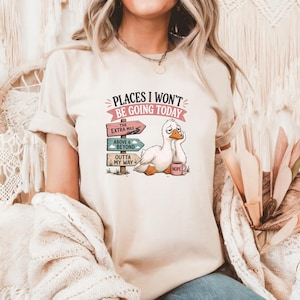 May include: A beige short-sleeve t-shirt featuring a graphic of a tired white duck holding a pink mug labeled NOPE. Next to the duck is a signpost with three signs reading THE EXTRA MILE, ABOVE & BEYOND, and OUTTA MY WAY, with the text PLACES I WON'T BE GOING TODAY.