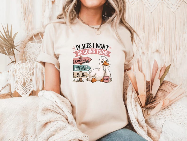 May include: A beige short-sleeve t-shirt featuring a graphic of a tired white duck holding a pink mug labeled NOPE. Next to the duck is a signpost with three signs reading THE EXTRA MILE, ABOVE & BEYOND, and OUTTA MY WAY, with the text PLACES I WON'T BE GOING TODAY.