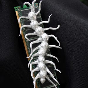 Puede incluir: Una escultura de ciempiés plateada y estilizada con múltiples patas, apoyada sobre un módulo de memoria de ordenador verde y dorado. La escultura tiene un cuerpo segmentado y está sobre un fondo negro.