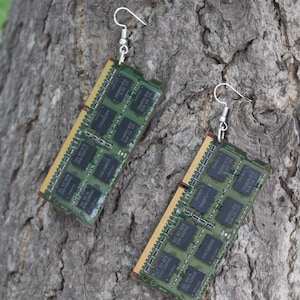 Op de afbeelding: Een paar unieke oorbellen gemaakt van hergebruikte computer RAM-chips. De oorbellen hebben zilverkleurige oorhaken en de groene en gouden printplaatcomponenten van de RAM-chips. De oorbellen worden getoond tegen een bruine, getextureerde achtergrond.