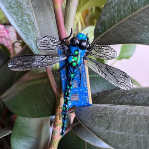 Puede incluir: Una escultura artesanal de libélula con cuerpo azul y verde, ojos negros y alas intrincadas. La libélula está montada sobre una placa de circuito azul, sobre un fondo de hojas verdes.