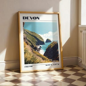 May include: A framed print of a coastal scene, with the word "DEVON" at the top. The image shows green cliffs, blue sea, and an island. The print also includes the text "ENGLAND" and geographical coordinates.