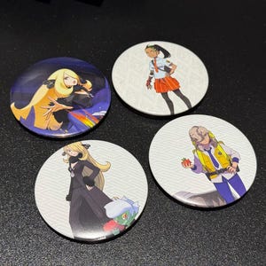 Puede incluir: Cuatro botones redondos con ilustraciones de personajes de estilo anime. Un botón presenta un personaje con cabello rubio largo sobre un fondo oscuro con un arcoíris. Los otros tres botones presentan diferentes personajes sobre un fondo blanco.