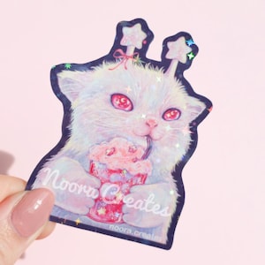 May include: A holographic sticker featuring a fluffy white cat with large pink eyes, wearing star-shaped antennae, and holding a pink drink with a straw. The sticker includes the text Noora Creates and noora.creates in white script lettering.