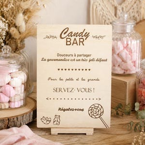 Op de afbeelding: Een rechthoekig bord van lichtgekleurd hout met donkerbruine gegraveerde tekst: Candy BAR, Douceurs à partager, La gourmandise est un très joli défaut, Pour les petits et les grands, SERVEZ-VOUS! en Régalez-vous, inclusief beer- en lolly-iconen.
