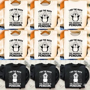 Op de afbeelding: Crème en zwarte sweatshirts met de tekst "I DID THE MATH THE ANSWER IS PENGUIN." Het ontwerp toont een cartoonpinguïn met een hoed en vlinderdas, omringd door wiskundige symbolen. De sweatshirts worden op houten hangers getoond.