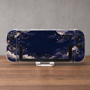 May include: A handheld gaming console featuring a dark blue skin with a celestial pattern of white clouds, golden stars, and crescent moons. The device is held in a black plastic stand and includes black buttons, joysticks, and directional pads.
