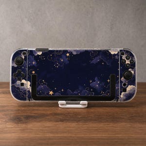 May include: A handheld gaming console featuring a dark blue skin with a celestial pattern of golden stars, crescent moons, and soft clouds. The device has black control sticks, directional buttons, and action buttons on both sides of the screen.
