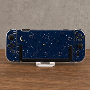May include: A handheld gaming console featuring a dark blue skin with a celestial pattern of gold stars, constellations, a crescent moon, and heart outlines. The device is encased in a clear protective shell and rests on a white plastic stand.