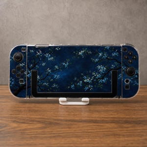 May include: A handheld gaming console featuring a dark blue skin with a pattern of light blue cherry blossom branches. The device has a clear protective case, black control sticks, and buttons on the left and right sides, resting on a white stand.