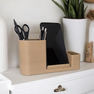 Könnte beinhalten: Ein beiger Schreibtisch-Organizer mit Fächern für Stifte, Scheren und ein Tablet. Der Organizer besteht aus einem strukturierten Material und steht auf einer weißen Oberfläche. Ein schwarzes Tablet befindet sich in der Mitte.
