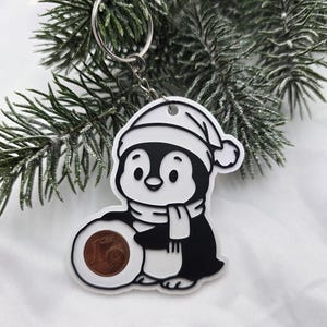 Könnte beinhalten: Ein schwarz-weißer Acryl-Schlüsselanhänger mit einem Cartoon-Pinguin, der eine Wintermütze und einen Schal trägt. Der Pinguin hält einen kreisförmigen Ausschnitt, der eine echte Ein-Euro-Cent-Münze enthält. Mit silbernem Metallring.