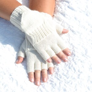 Może przedstawiać: Kremowe rękawiczki bez palców wykonane z dzianiny. Rękawiczki mają prążkowany mankiet i guzik. Prezentowane są na białej, teksturowanej powierzchni. Rękawiczki mają za zadanie ogrzewać dłonie, pozostawiając palce wolne.