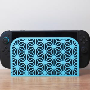 May include: A light blue geometric patterned stand for a black gaming console. The stand is made of a laser-cut material and features a repeating starburst design. The console is black with blue and black controls.