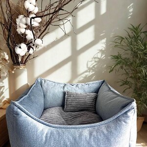 May include: A light blue denim pet bed with a rectangular shape and a soft, cushioned interior. The bed includes two gray pillows for added comfort. The bed is designed for small to medium-sized pets.