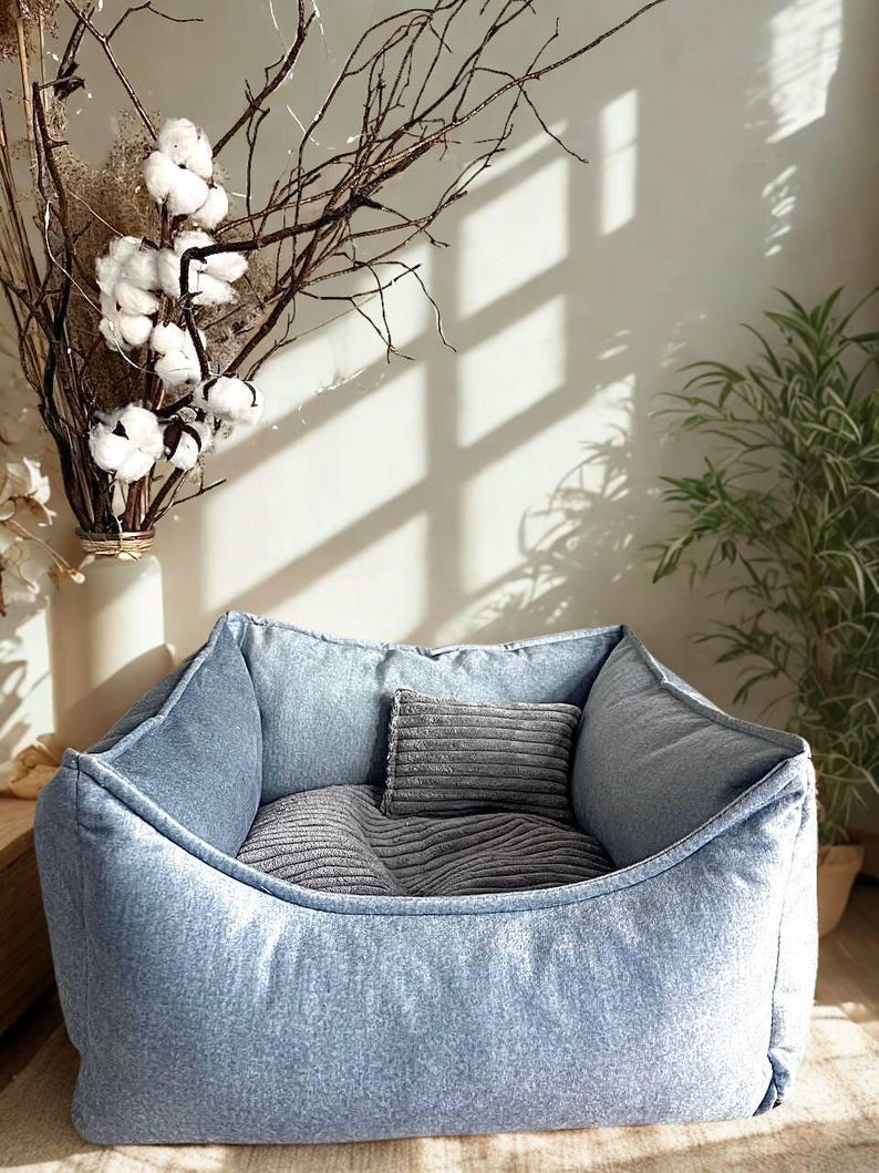 May include: A light blue denim pet bed with a rectangular shape and a soft, cushioned interior. The bed includes two gray pillows for added comfort. The bed is designed for small to medium-sized pets.