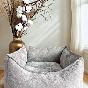 May include: A gray pet bed with a textured fabric design. The bed has a square shape with raised sides and a cushioned interior. A gold vase with dried branches and cotton flowers is in the background.