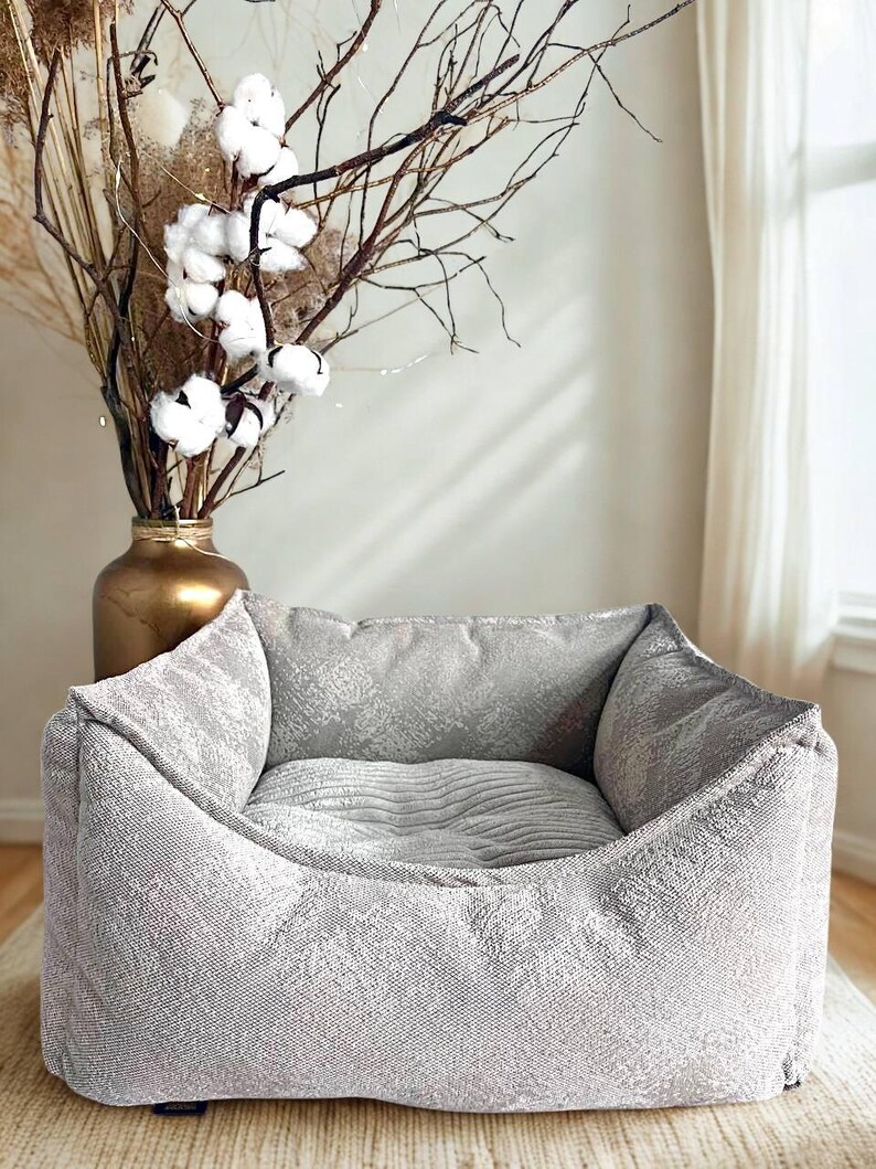 May include: A gray pet bed with a textured fabric design. The bed has a square shape with raised sides and a cushioned interior. A gold vase with dried branches and cotton flowers is in the background.