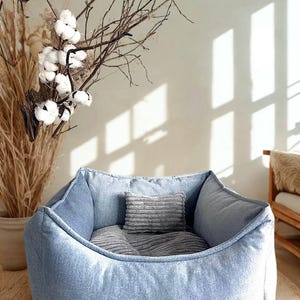May include: A light blue denim pet bed with a soft, rounded shape. The bed contains two gray pillows and is designed for comfort. The background includes dried plants and a neutral-toned wall, creating a cozy aesthetic.