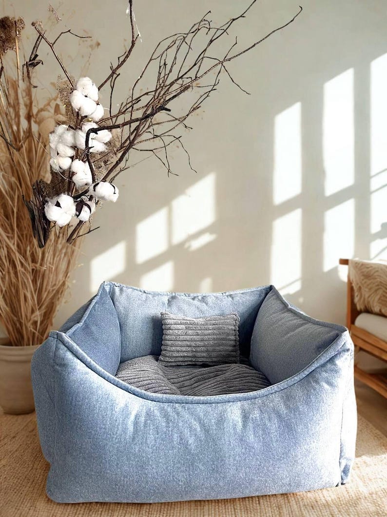 May include: A light blue denim pet bed with a soft, rounded shape. The bed contains two gray pillows and is designed for comfort. The background includes dried plants and a neutral-toned wall, creating a cozy aesthetic.