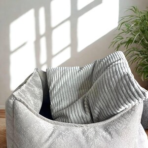 May include: A gray pet bed with a textured exterior and a rectangular shape. Inside the bed are two gray corduroy pillows. The bed is placed on a light brown surface, with a plant and window shadows in the background.