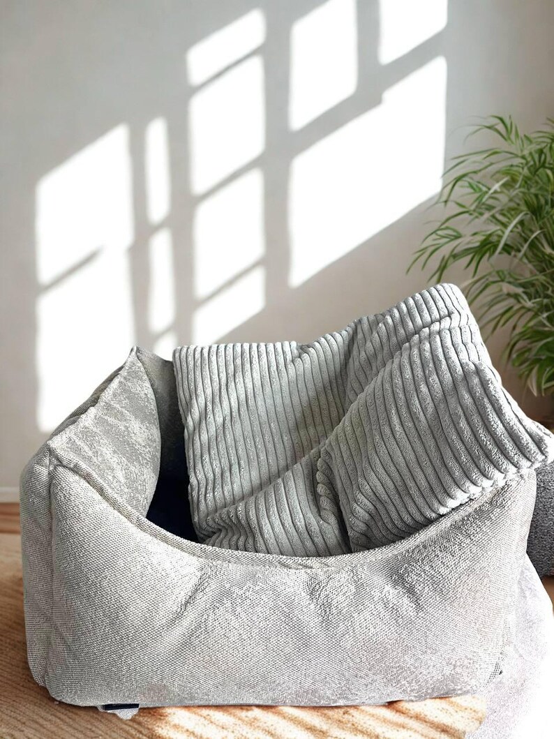 May include: A gray pet bed with a textured exterior and a rectangular shape. Inside the bed are two gray corduroy pillows. The bed is placed on a light brown surface, with a plant and window shadows in the background.