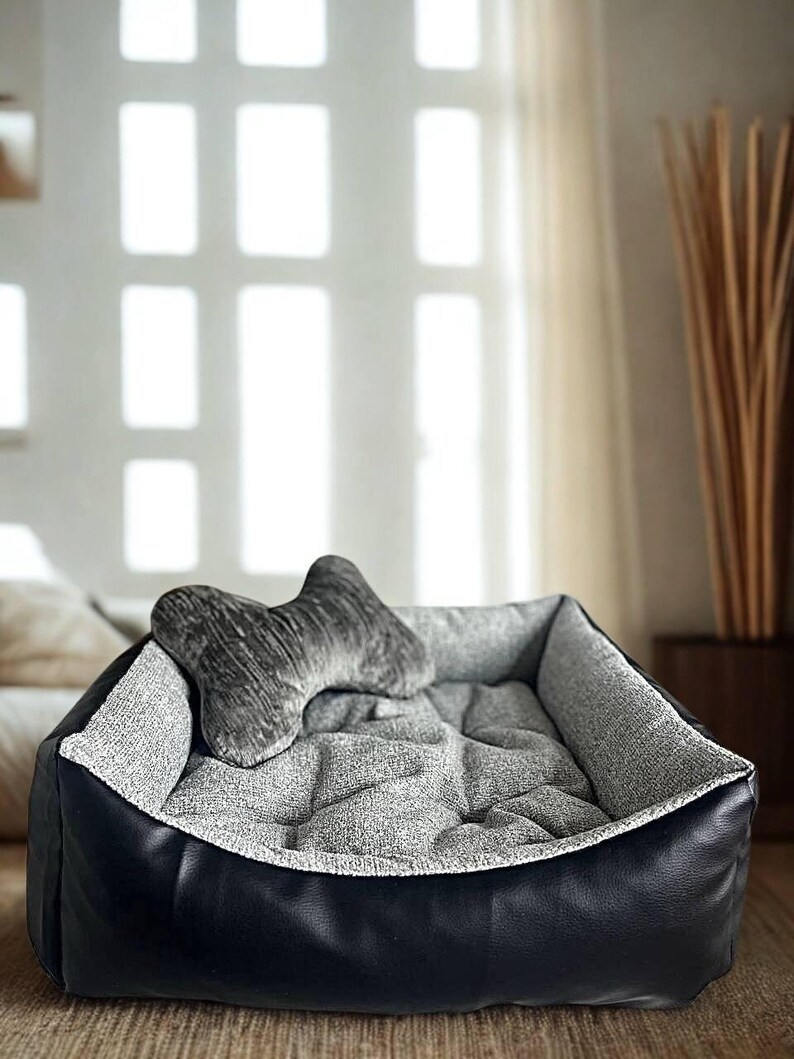 May include: A dog bed with a black faux leather exterior and a gray fabric interior. A bone-shaped pillow rests inside the bed. The bed is designed for comfort and style, suitable for small to medium-sized dogs.