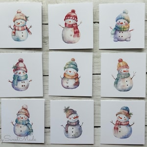 May include: Nine square greeting cards featuring watercolor illustrations of snowmen. Each snowman wears a different colored hat and scarf. The snowmen have twig arms and button eyes. The cards are arranged on a white wooden surface.