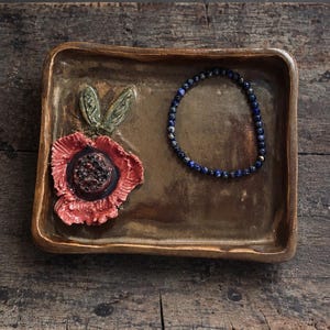Puede incluir: Una bandeja rectangular de cerámica marrón con una flor roja esculpida con hojas verdes en el lado izquierdo y una pulsera circular de cuentas azules sobre el lado derecho de la superficie lisa y esmaltada.