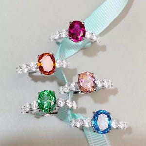 Könnte beinhalten: Eine Auswahl an Silberringen, jeweils mit einem ovalen Edelstein in verschiedenen Farben: Rubinrot, Orange, Rosa, Grün und Blau. Jeder Ring hat kleinere, klare Steine am Band. Die Ringe werden auf einem hellblauen Band präsentiert.