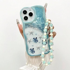Può includere: Una custodia trasparente per telefono con un design a tema oceano blu e bianco. La custodia presenta un motivo a onde, stelle marine e tartarughe marine. È attaccato un ciondolo per telefono con perline con accenti di stelle e perle.