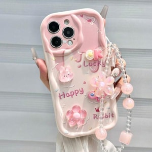 Può includere: Custodia per telefono rosa con design a coniglio e fiori 3D. La custodia presenta le parole "Happy" e "Rabbit" stampate, insieme a un cinturino di perline. Progettata per uno smartphone.