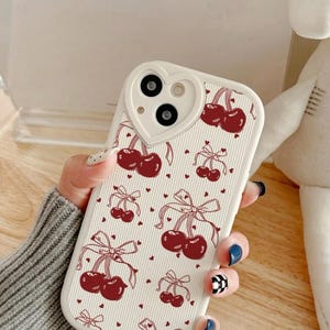 Op de afbeelding: Een witte telefoonhoes met een hartvormige camera-uitsparing. De hoes heeft een herhalend patroon van rode kersen met strikken en kleine rode hartjes. De hoes wordt vastgehouden in een hand met gelakte nagels.