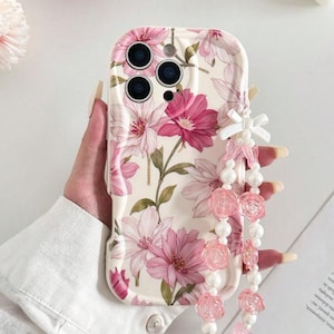 Puede incluir: Funda de teléfono floral con flores rosas y blancas sobre un fondo crema. Tiene una correa de cuentas a juego con detalles de rosas rosas y perlas blancas. La funda del teléfono es sostenida por una persona.