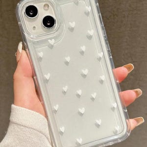 May include: Transparent phone case featuring a white heart pattern. The case has a clear design with raised white hearts across the back. The phone case is held in a hand.
