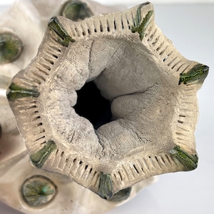 May include: A handcrafted ceramic vase with a unique, textured design. The vase features a light beige color with green accents and a star-shaped opening. The interior is a dark, cavernous space.