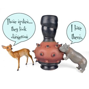 May include: A ceramic vase with a dark brown top and a reddish-brown body adorned with black spikes. A fawn and a hippo toy are positioned near the vase, with speech bubbles that read "Those spikes... they look dangerous" and "I love them."