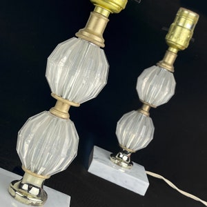 May include: Two vintage table lamps with clear, faceted glass bodies and brass-toned metal accents. The lamps sit on square, white marble bases. The lamps have a classic, elegant design, perfect for a bedroom or living room.