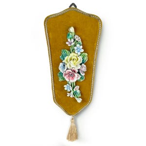 May include: Mustard-colored velvet wall hanging with a floral ceramic arrangement. The arrangement features a yellow rose, pink and blue flowers, and green leaves. A gold trim and tassel complete the design.