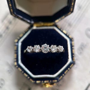 May include: A silver-toned ring featuring five round-cut clear stones set in a row, with the center stone being the largest. The ring is displayed inside an open, dark blue, octagonal-shaped jewelry box with a dark blue velvet interior lining.