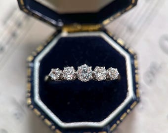 Könnte beinhalten: Ein silberfarbener Ring mit fünf runden, klaren Steinen in einer Reihe, wobei der mittlere Stein am größten ist. Der Ring befindet sich in einer geöffneten, dunkelblauen, achteckigen Schmuckschatulle mit dunkelblauem Samtfutter.