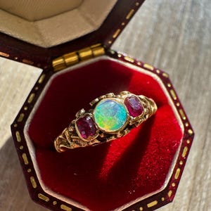 May include: A gold ring featuring a central round cabochon opal displaying iridescent flashes of green and blue, flanked by two rectangular red rubies. The ring has an ornate, textured metal band and is presented inside a dark red velvet box.